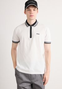 Vitt polo-shirt med svart dragkedja och krage, med grå accentdetaljer. Bärs med grå byxor och en svart keps som har "BOSS" på.