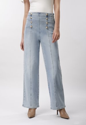 Svetlomodré vysokopásové denimové nohavice s dekoratívnymi gombíkmi, v kombinácii s béžovými lodičkami so špicatou špičkou a bielym tielkom bez rukávov.