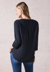 Marineblauwe blouse met een zachte textuur, driekwartmouwen en plooidetail aan de achterkant. Draagt zich met blauwe jeans, wat een casual stijl laat zien.