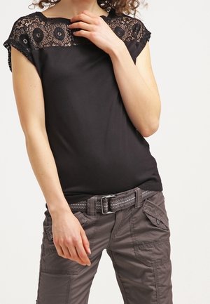 Vrouw draagt een zwart topje met kanten accenten en grijze cargobroek met een geweven riem, poseert met één hand bij haar nek.