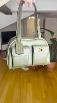 Mala de mão em pele verde com dois bolsos frontais, ferragens douradas, alças duplas e uma etiqueta. Textura lisa com design estruturado.