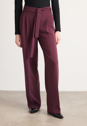 Pantalones de pierna ancha burdeos con un cinturón de lazo, pliegues y tela suave. Combinados con una blusa negra de manga larga y zapatos negros de punta.