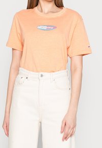 Persikofärgad bomulls-t-shirt med rund hals, korta ärmar och en oval logotypgrafik i blått och rosa på bröstet. Matchas med vita jeans.