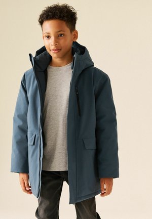 Ragazzo che indossa un cappotto invernale blu con cappuccio sopra una camicia grigia e pantaloni neri, in piedi davanti a uno sfondo chiaro e uniforme.