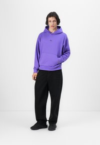 Lila Kapuzenpulli mit einer Fronttasche, gerippten Bündchen und Saum, kombiniert mit schwarzen weit geschnittenen Hosen und schwarzen Turnschuhen, vor einem weißen Hintergrund präsentiert.