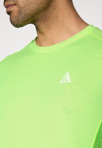 Αθλητική μπλούζα σε νέον πράσινο με ομαλή υφή, με λογότυπο σε λευκό χρώμα στο σχήμα ενός ανεστραμμένου τριγώνου στο στήθος.
