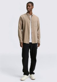 Hellbraunes, geripptes Hemd mit Knöpfen und Brusttasche, getragen über einem weißen T-Shirt und kombiniert mit schwarzen Hosen und weißen Sneakers.