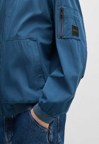 Blau leichte Jacke mit einer Reißverschlusstasche am linken Ärmel, gerippten Bündchen und glatter Textur. Über leicht ausgebleichter Jeans getragen.