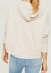 Beżowy skrócony hoodie z kapturem na sznurki, elastycznym dołem i długimi rękawami, wykonany z miękkiego materiału, noszony na jasnoniebieskich dżinsach.