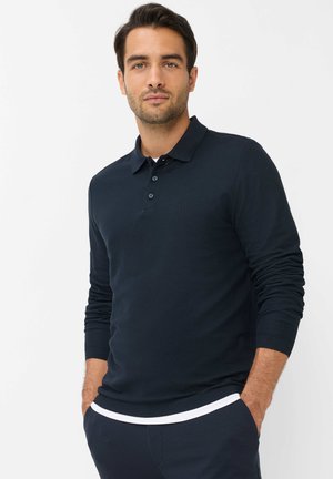Man draagt een donkerblauw langarm poloshirt met handen in de zakken, staand tegen een effen witte achtergrond.