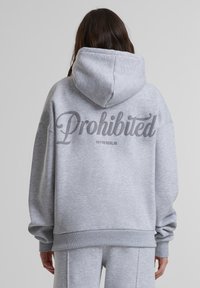 Pilkas džemperis su minkšta, tekstūruota paviršiumi. Ant nugaros yra didelė tamsiai pilka „Draudžiama“ grafika, o po ja – „10119 BERLYNAS“.}}