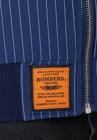 Tessuto blu a righe sottili con cerniera argento e etichetta rettangolare arancione con scritto "Bombers Original U.S. Air Force flight jacket."