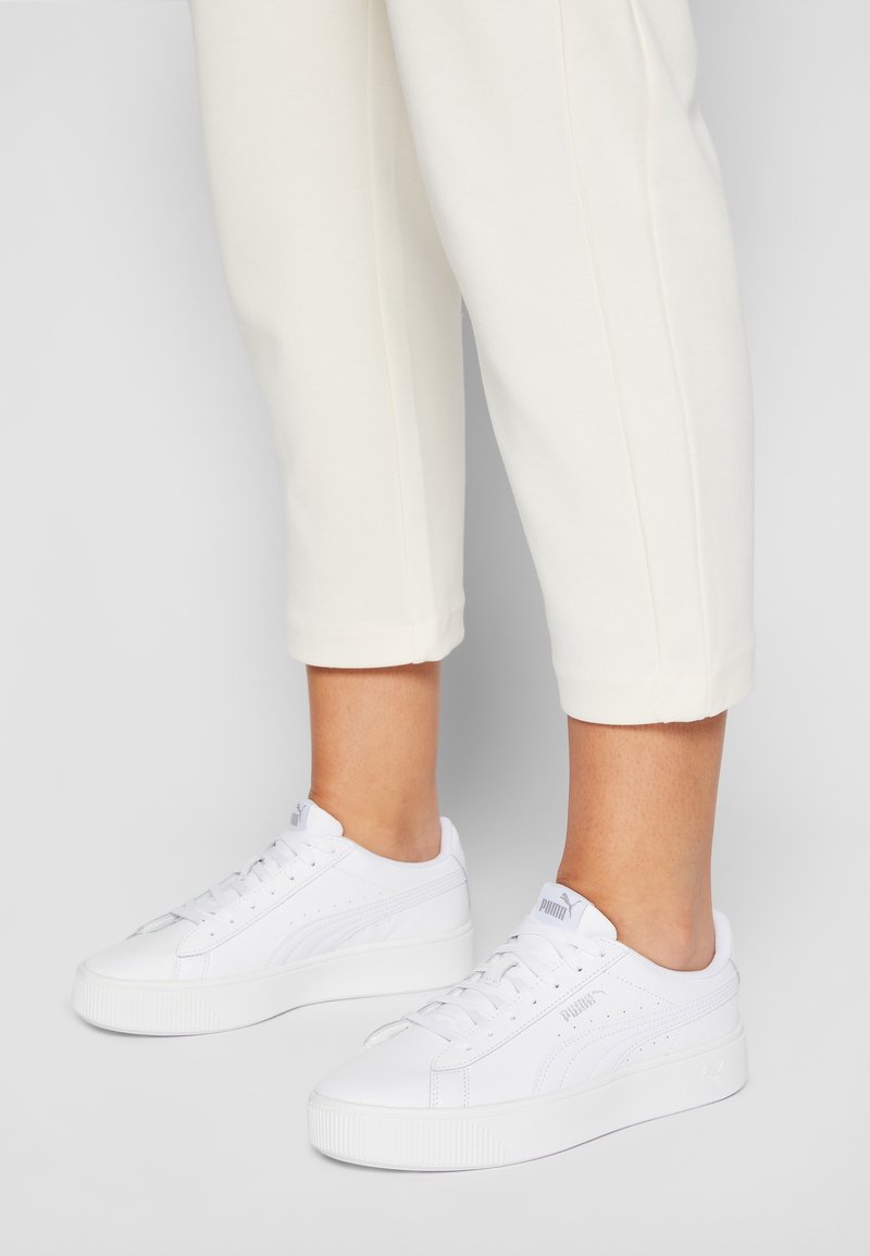 Baskets en cuir blanc avec une semelle épaisse, dotées de détails perforés, de lacets plats et d'un petit logo sur le côté. Portées avec un pantalon court crème.