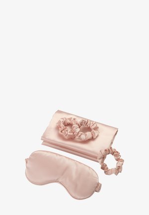 Set da notte in raso rosa chiaro con federa piegata, maschera per occhi coordinata e tre elastici per capelli scrunchie disposti su sfondo bianco.