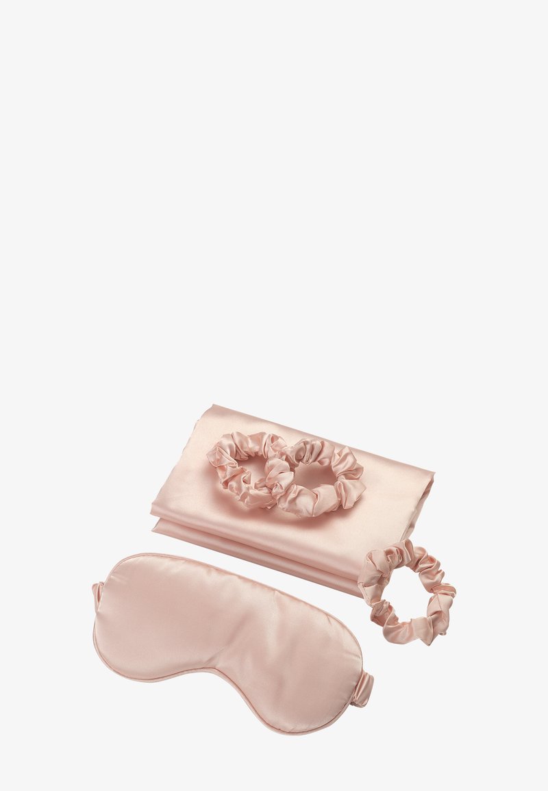 Ensemble de nuit en satin rose clair avec une taie d'oreiller pliée, un masque pour les yeux assorti et trois chouchous disposés sur un fond blanc.