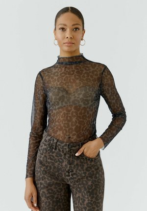 Frau trägt transparentes langärmliges Leopardenprint-Top über passendem Bralette und hoch taillierter Leopardenprint-Jeans mit der Hand in der Tasche.