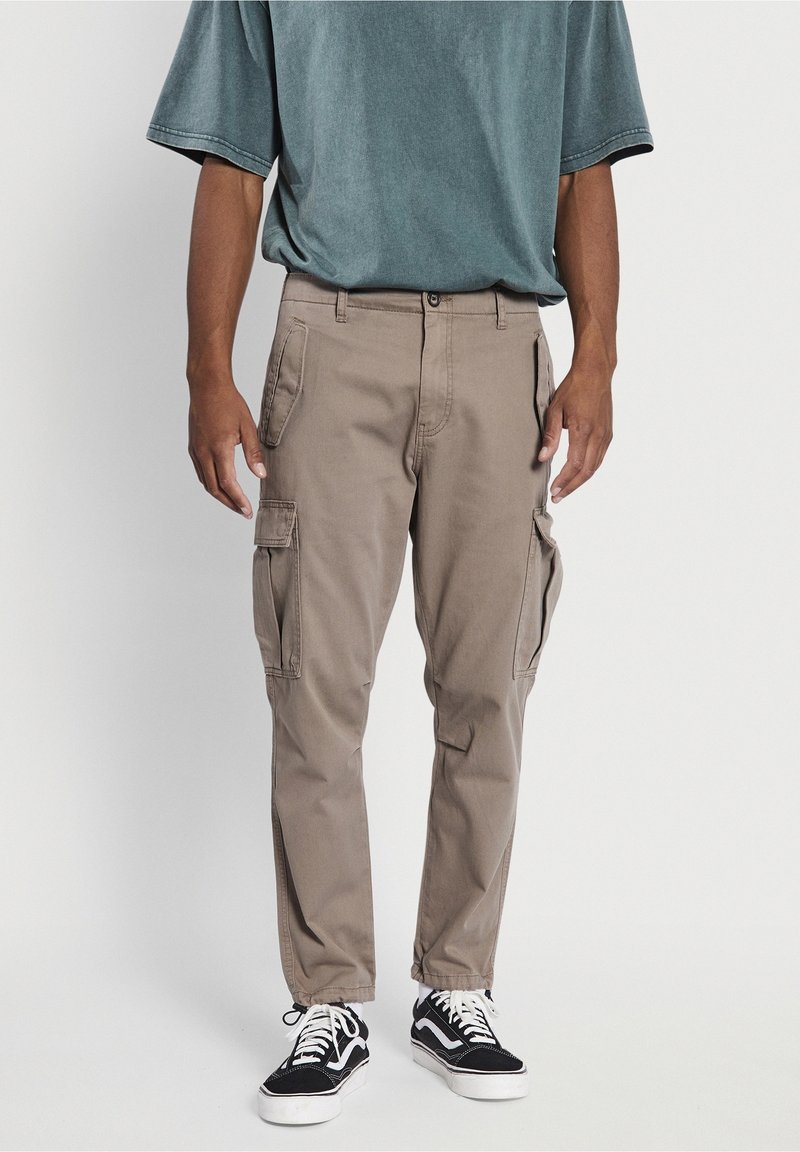 Homme portant un pantalon cargo beige avec des poches latérales, un t-shirt bleu-gris ample et des baskets noires et blanches, debout devant un fond uni.