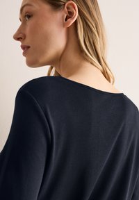 Top in maglina blu navy con scollatura a barca, caratterizzato da una texture liscia e una vestibilità morbida. I dettagli includono una cucitura sottile lungo la spalla.