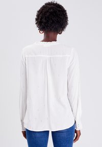 White long-sleeve blouse in lightweight fabric, featuring subtle vertical shimmer patterns, a gathered neckline, and buttoned cuffs.