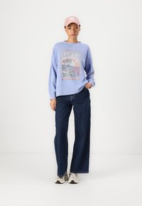Lichtblauw oversized sweatshirt met een grafische print, gecombineerd met wijde donker denim jeans en een roze pet. Sneakers maken de outfit compleet.