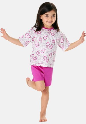 Smiling young girl balancing on one foot with arms outstretched, wearing a white shirt with pink smiley faces and pink shorts.