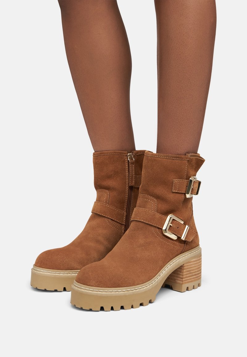 Botines de ante marrón con dos correas con hebilla, una cremallera lateral y un tacón plataforma grueso y dentado. Textura suave y diseño robusto.