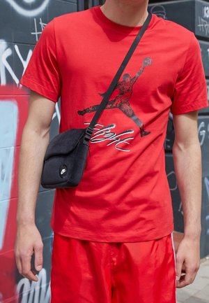 Personne portant un t-shirt rouge avec un graphisme noir de joueur de basketball et un short rouge, portant un petit sac bandoulière noir Nike dans un décor urbain avec graffiti.