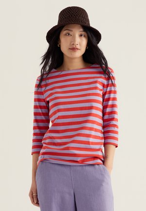 Nő hosszú fekete hajjal, barna horgolt sapkát viselve, piros-lila csíkos háromnegyedes ujjú felsőben és világoslila nadrágban, egyik kezével a zsebében.