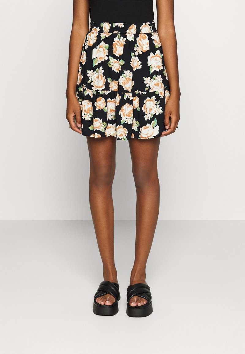 Personne portant une mini-jupe noire à fleurs beiges et blanches, un haut noir, et des sandales à plateforme noires, debout devant un fond uni.