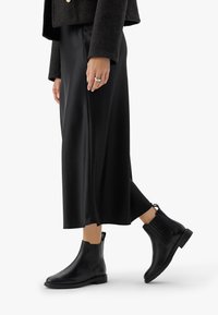 Sieviete, kura valkā melnu midi svārku, tumšu īsu jakas, melnus ādas potīšu zābakus un sudraba gredzenu uz kreisās rokas.