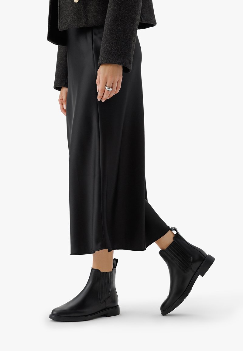 Sieviete, kura valkā melnu midi svārku, tumšu īsu jakas, melnus ādas potīšu zābakus un sudraba gredzenu uz kreisās rokas.