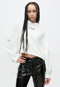 Balta nostriķēta kapuce ar uzrakstu "PINKO" priekšpusē, kombinēta ar melnām ādas imitācijas biksēm ar spīdīgu virsmu un piegulošu vidukli.