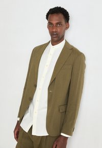 Olive green linen blazer with a single button and a notch lapel, worn over a white shirt with a stand-up collar. Smooth texture.