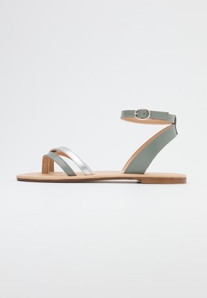 Sandal en cuir vert avec des brides métalliques argentées, design plat, bout ouvert, boucle à la cheville et semelle en bois naturel. Style simple et minimaliste.