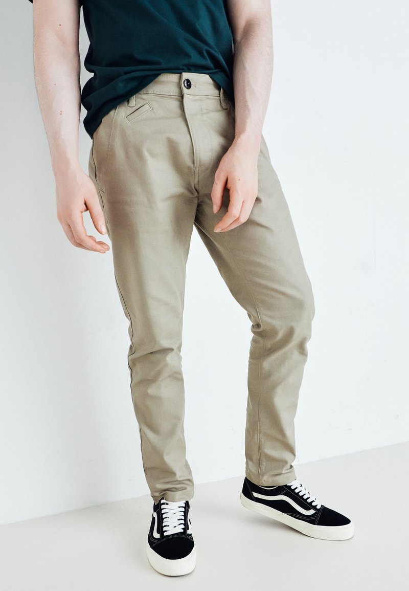 Personne portant un pantalon beige slim et des baskets noir et blanc, debout devant un fond blanc uni.