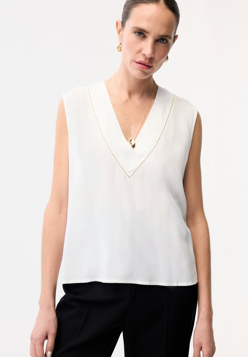 Mujer con blusa blanca sin mangas de escote en V, pantalones negros, pendientes dorados colgantes y collar con colgante dorado sobre fondo liso.