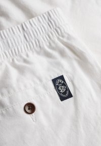 Weiße Stoffshorts mit einem elastischen Bund, ausgestattet mit einem braunen Knopf und einem blauen Etikett mit der Aufschrift "Super Dry." Strukturierter Baumwollstoff.
