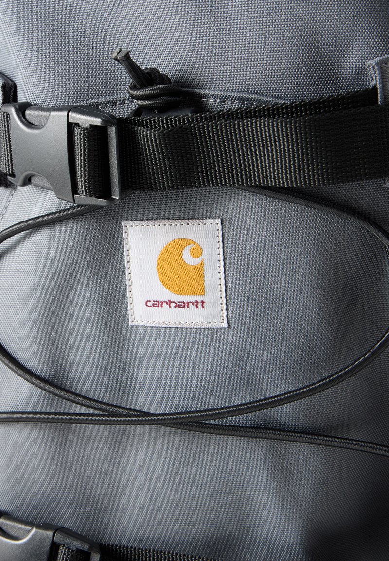 Grauer Stoffrucksackabschnitt mit schwarzen verstellbaren Gurten und einem weißen Aufnäher mit gelbem Carhartt-Logo und rotem Text.