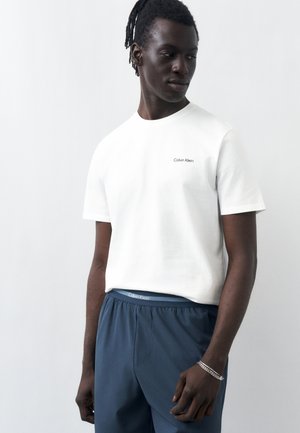 Férfi, fehér Calvin Klein pólót és kék nadrágot visel, balra lefelé néz, egyszerű világos háttér előtt.