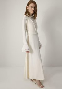 Lang witte jurk met een hoge halslijn, uitlopende mouwen en een diepe V-hals. Gladde stof met een vloeiende silhouet, subtiele zijsplitsingen en minimale details.