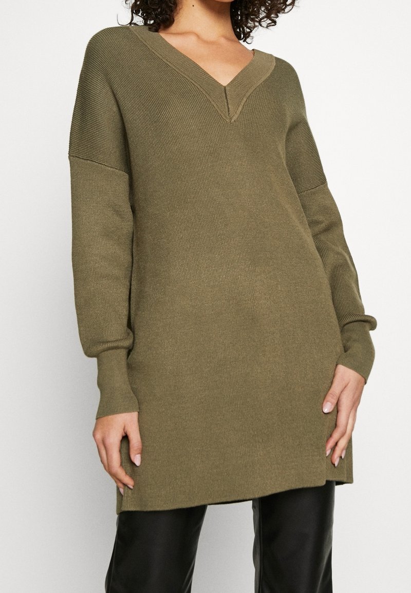 Robe tricotée vert olive avec un décolleté en V, des épaules tombantes et une texture côtelée. Elle présente des manches longues et des fentes latérales au bas.