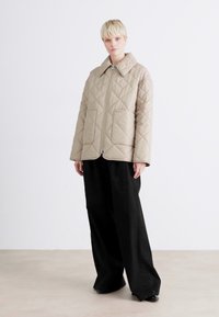 Μπεζ επενδυμένο μπουφάν με γιακά, εμπρός φερμουάρ και διαμαντέ σχέδιο. Συνδυασμένο με φαρδιά μαύρα παντελόνια, μπροστά σε ουδέτερο φόντο.