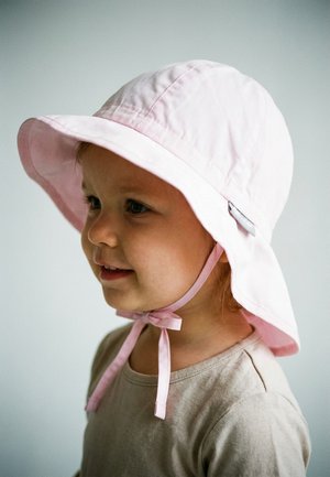 Young child smiling, wearing a light pink sun hat with chin straps tied and a beige shirt, against a neutral background.