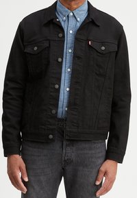 Black denim jacket featuring a button-up front, two chest pockets, and a collar. Worn over a light blue button-down shirt.