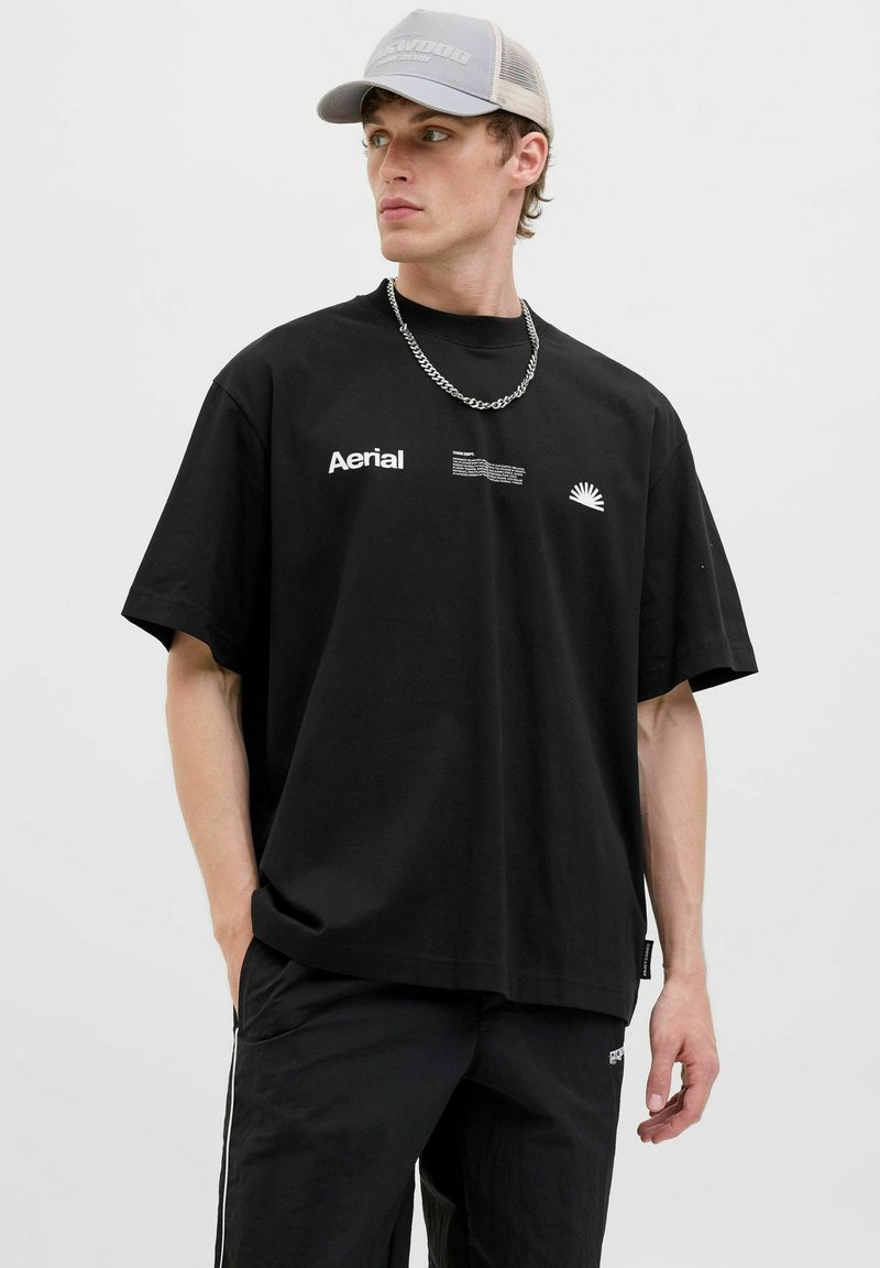 Camiseta negra de gran tamaño con mangas cortas, que presenta un texto blanco y un gráfico de sol. Combinada con pantalones negros y una gorra gris clara con malla.
