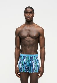 Homme musclé à la peau foncée debout, portant un boxer bleu et vert à motifs avec une ceinture bleu clair, sur un fond gris uni.