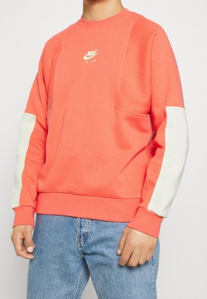 Mężczyzna ubrany w koralową bluzę Nike Air z białymi panelami na rękawach i jasnoniebieskie dżinsy, stojący na jednolitym jasnym tle.