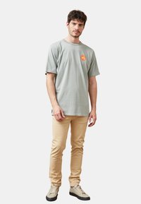 Camiseta de algodón gris claro con manga corta y escote redondo, que presenta un logo en forma de círculo rojo. Combinada con pantalones beige ajustados y zapatillas gris.