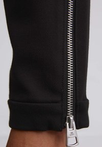 Zwarte stoffen broek met een zilveren metalen rits langs de zijkant, voorzien van een omgeslagen zoom voor een nette afwerking.