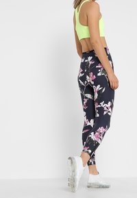 Neon zelený sportovní top, černé legíny s květinovým vzorem v růžové, bílé a zelené barvě a průhledné běžecké boty. Pohled zezadu, štíhlý střih.